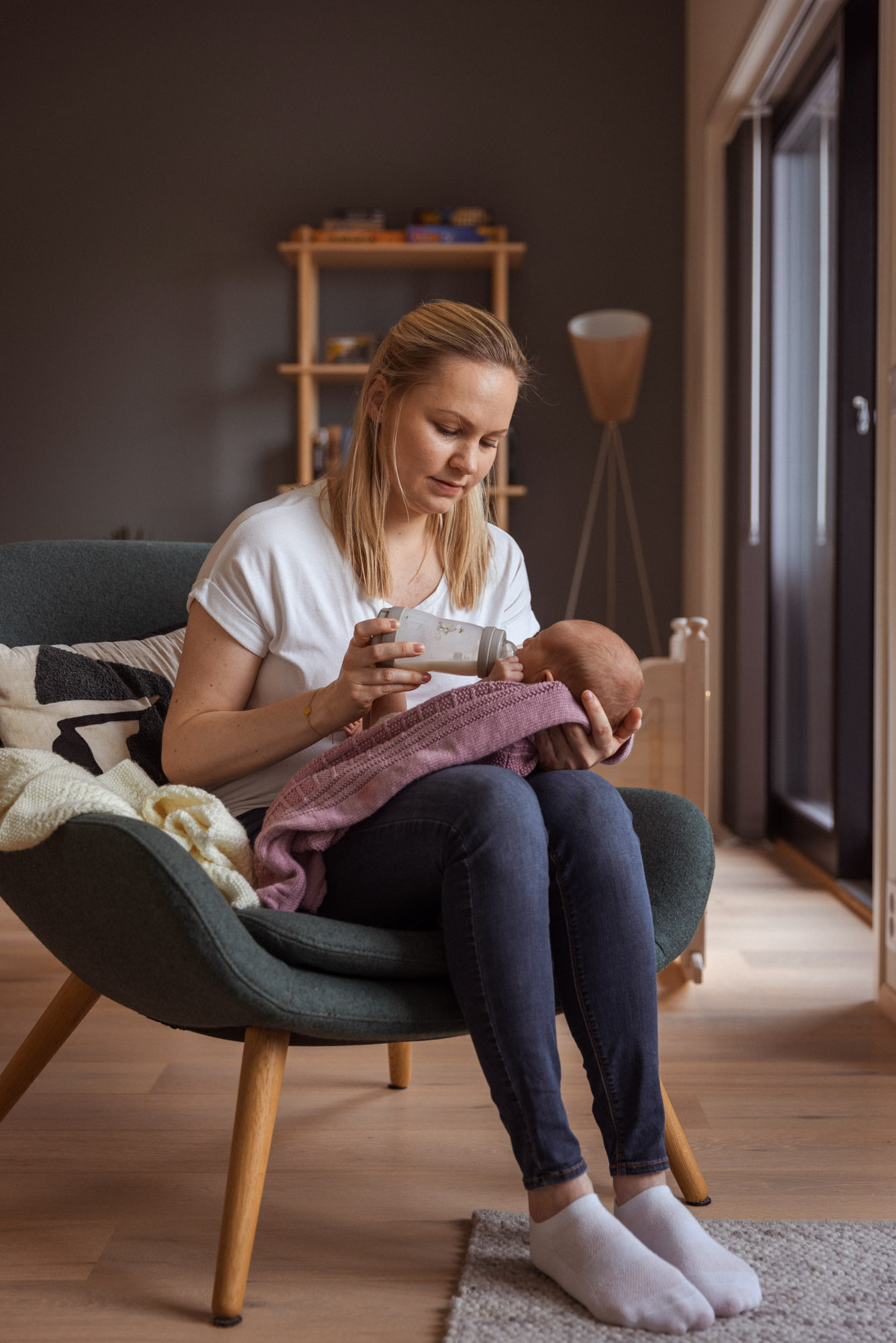 Livsstilsfoto av en mor som sitter i en lenestol og gir melk på flaske til sitt nyfødte barn. Babyen er inntullet i et rosa strikket teppe.