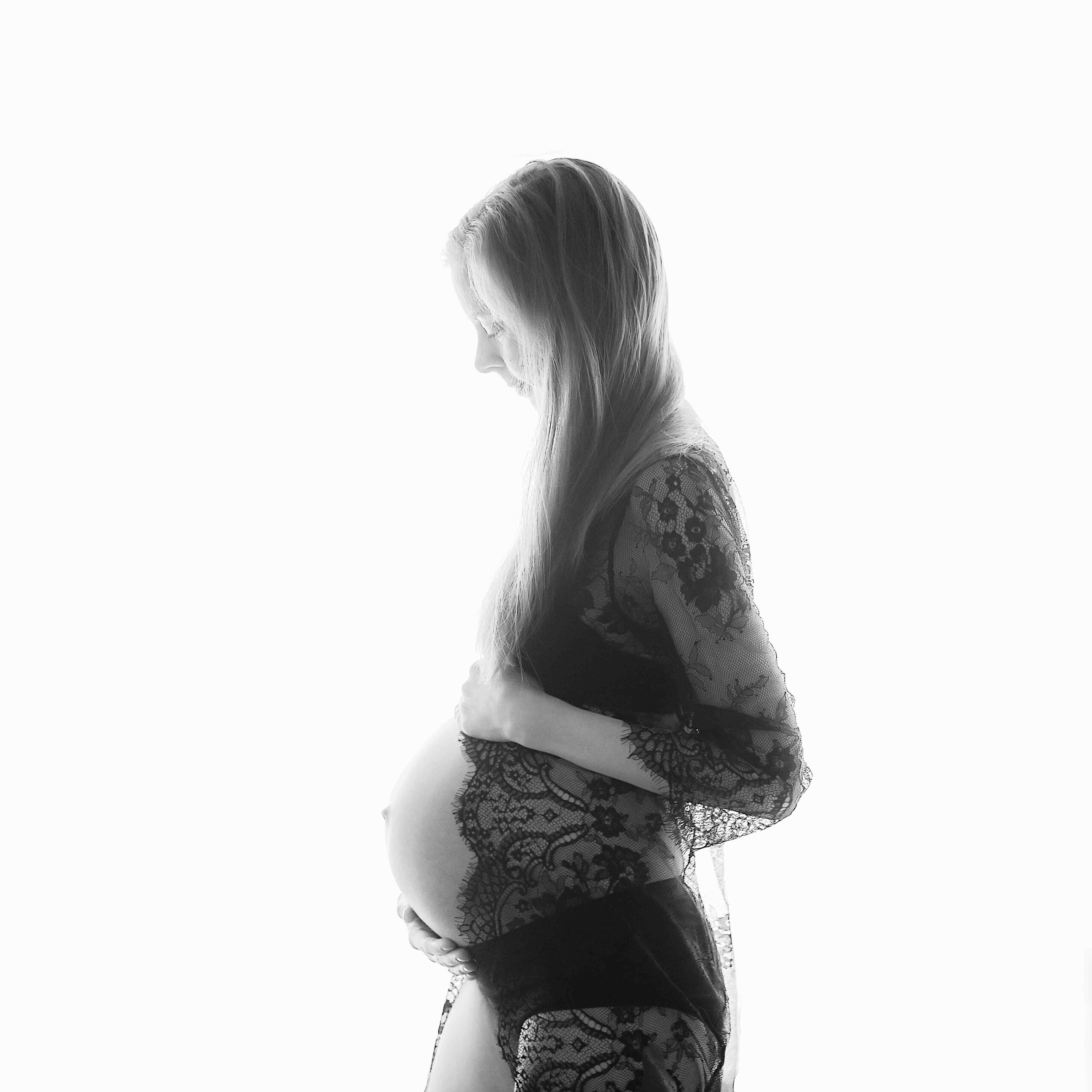 Sort/hvitt bilde av en gravid kvinne fotografert i profil. Sort blondetopp, langt hår, mor holder rundt magen sin.