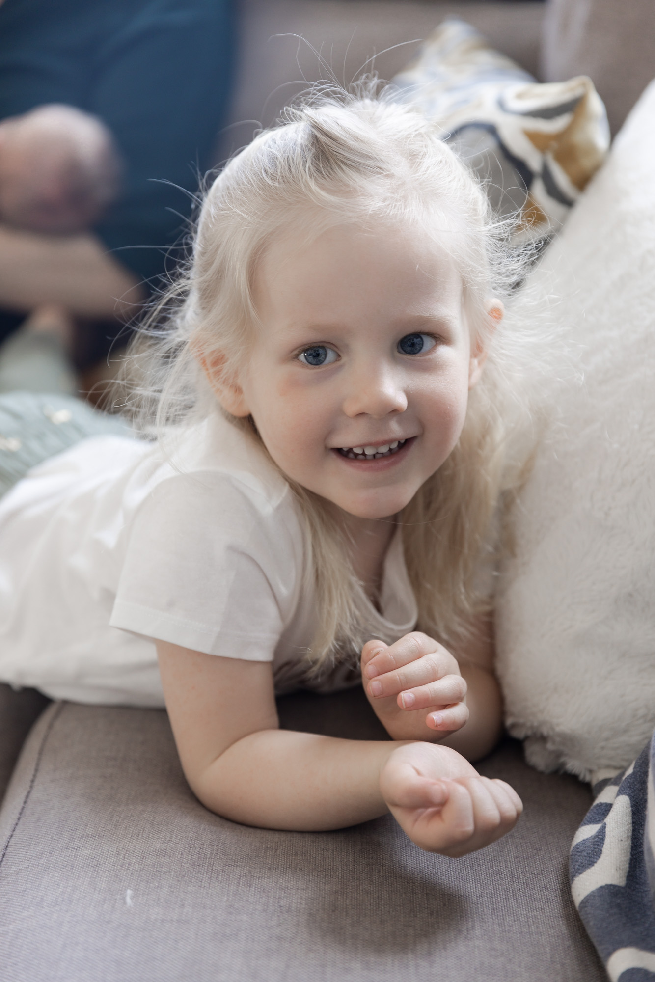 En treårig jente i hvit t-skjorte ligger på magen på en grå sofa og smiler mot kamera.