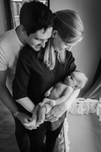 Sort/hvitt bilde av foreldre med baby; mor holder baby i armene sine, far står bak mor og omfavner henne og ser på babyen. 