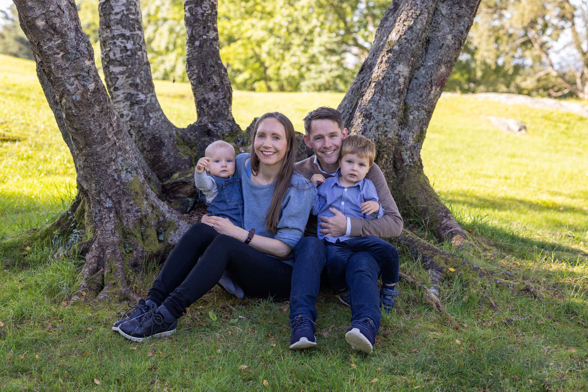 Familieportrett av to voksne og to barn som sitter inntil en klynge trær i Lynghaugparken i Bergen. Alle har noe blått eller brunt på seg. Sko til foto.