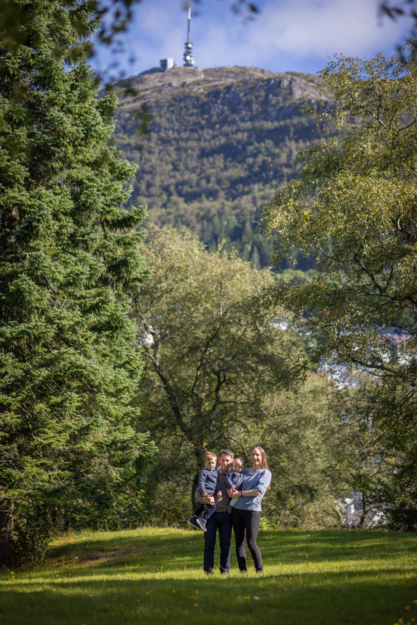 Familie på fire; to voksne og to små barn, fotografert i Langhaugparken i Bergen. Ulriken fjellet i bakgrunnen, familien står på en grønn plen med høye grønne trær rundt dem.