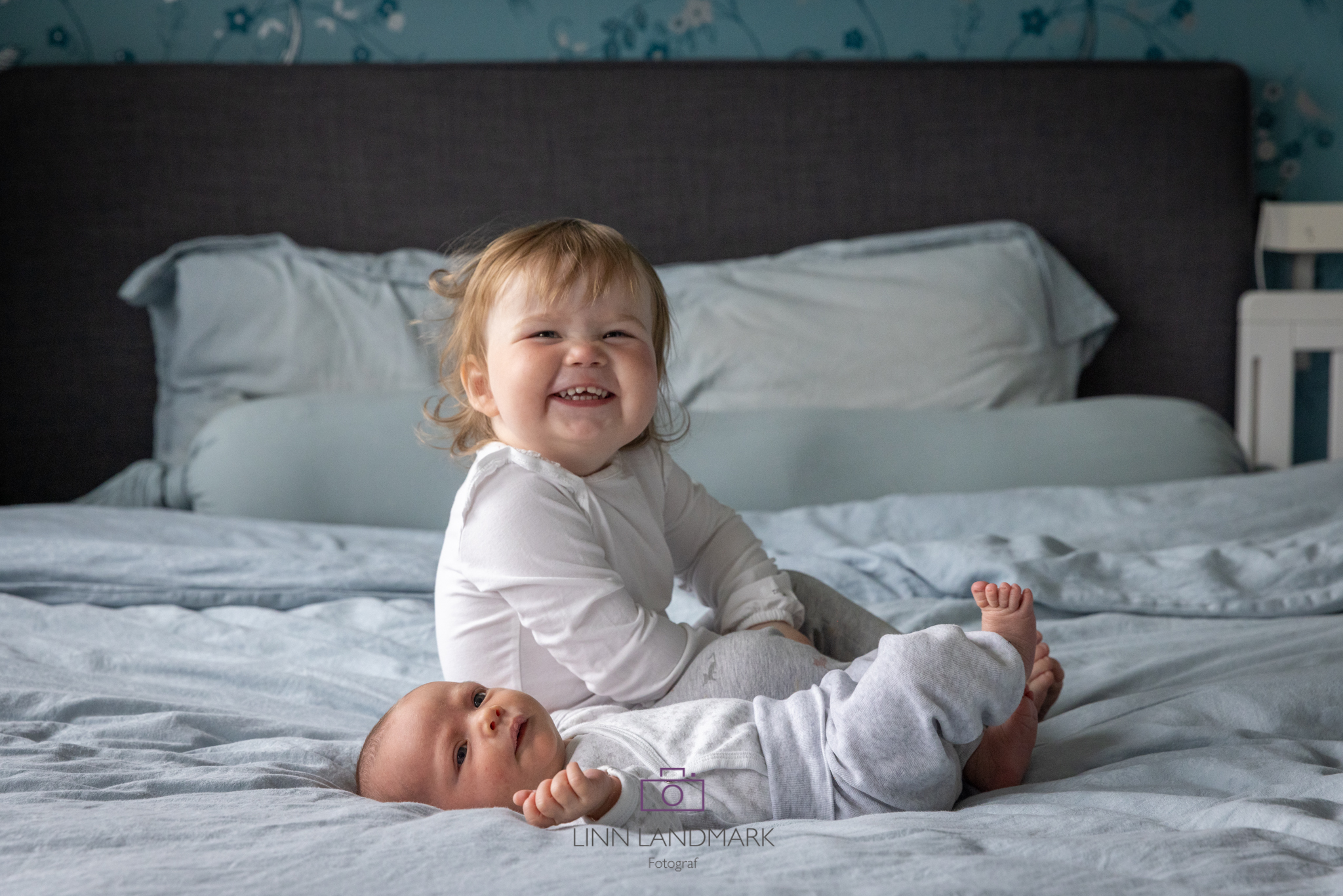 En nyfødt baby ligger på ryggen på en stor seng, storesøster på ett år sitter bak og smiler mot kamera. Blått sengetøy og puter, barna har hvit og grå klær på seg.