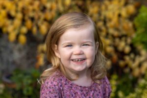 En treåring i lilla kjole smiler mot kamera. Gult høstløv i bakgrunnen. Fotografert av Fotograf Linn Landmark -Ta gode bilder av barna dine