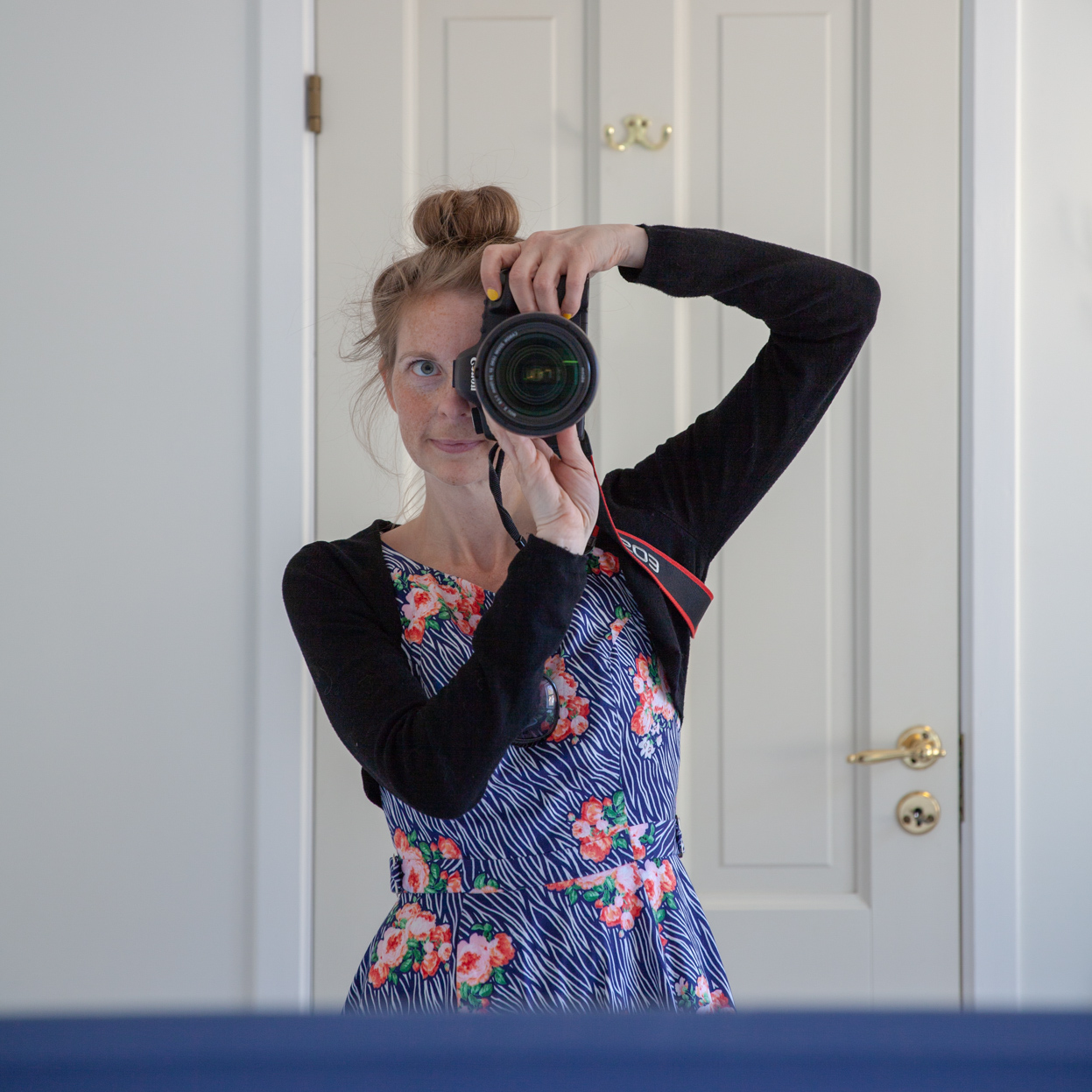 Selvportrett av Fotograf Linn Landmark som fotograferer seg selv mot et speil. Hun har donut i håret, sort bolero og blå blomstrete kjole. Hvit vegg og hvit dør i bakgrunnen.
