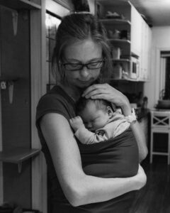 Sort/hvitt foto av en mor som holder sitt nyfødte barn i en bæretube. Babyen sover, mor holder en hånd på babyens hode. Babyutstyr du trenger.