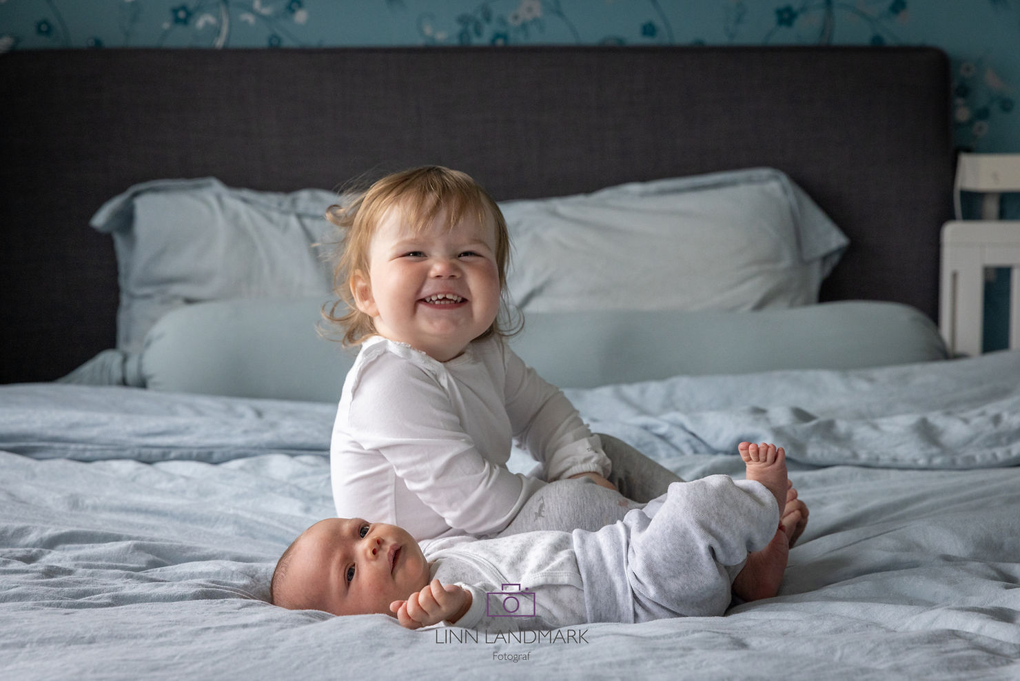En baby ligger på sengen og et småbarn sitter bak og smiler mot kamera. Søskenfoto av Fotograf Linn Landmark i Bergen. 