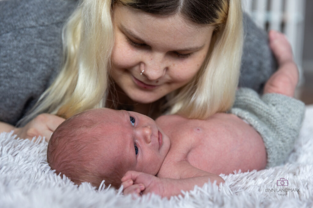 Bilde av en nyfødt baby som ligger på et grått pledd, mor lener seg over ham og ser på ham. Livsstilsfoto nyfødt hjemmefotografering i Bergen
