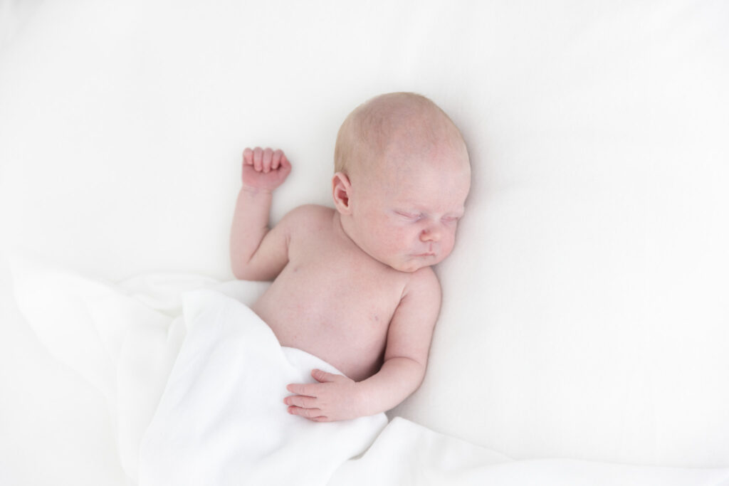 nyfødtfotografering i studio - en baby ligger på et hvitt pledd, med et annet hvitt pledd over nedre delen av kroppen, fra navlen og ned. Fotografert ovenfra, barnets ansikt er snudd mot lyset - hen ligger på ryggen.