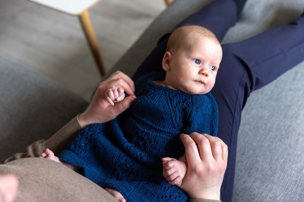 En baby ligger på mors fang under en hjemmefotografering i Bergen. Bildet er tatt ovenfra og ned, der man ser babyen ligge på fanget i mors armer - mor holder babyens hender, man ser deler av mors overkropp. Babyen ser til siden mot lyset og har på seg en blå strikket kjole.