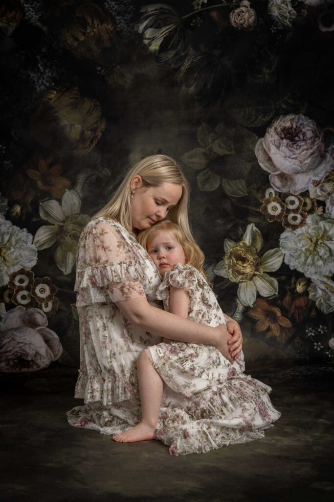 mamma og meg fotografering i Studio Landmark, Bergen. En mor sitter på kne og holder rundt sitt lille barn som sitter på fanget hennes og lener seg inn til mors bryst. begge har på blomsterkjoler. Mørk blomsterbakgrunn.