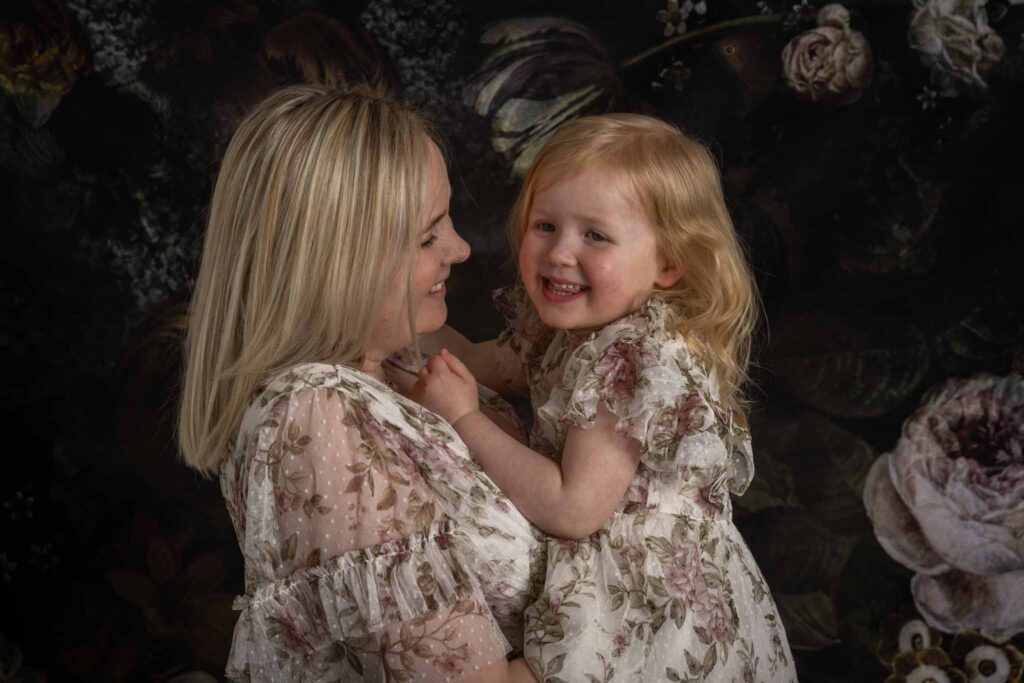 Mamma og meg fotografering i Bergen. En mor holder sin treårige datter i armene sine og ler mot henne, datteren ler tilbake. Utsnitt fotografert fra overarm til mor og opp. Begge er iført pen lys blomsterkjole i tyll.